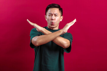 A young Asian man in a green T-shirt makes an "X" gesture with his arms crossed in front of him, accompanied by a disapproving expression, standing against a bold red background