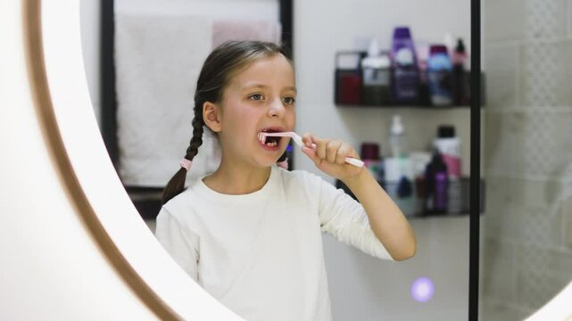 Portrait of young Caucasian girl with missing teeth brushing teeth while looking in bathroom mirror, emphasizing good oral hygiene habits.