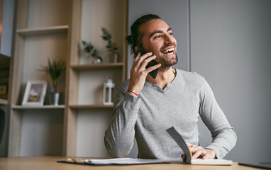 A cheerful man having a phone conversation while working from home, seated at a desk with a notepad.