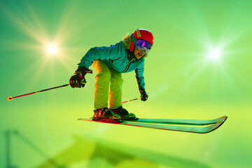 Low angle, side view of female athlete, skier in colorful gear mid-air jump neon green background....