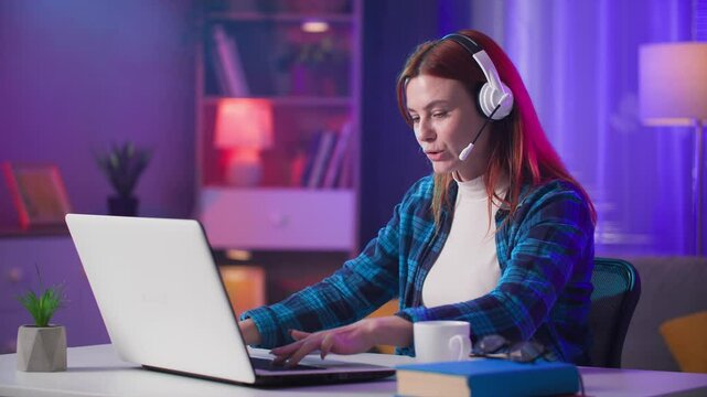 young woman with headset talking on video call with friends while playing video games together online on laptop computer sitting in room in evening