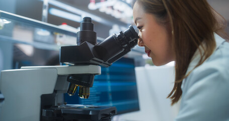 Medical Science Laboratory: Japanese Female Microbiologist Looking Through the Microscope, Analyzing Sample Genes and Writing Down Results on a Computer. Skillful Scientist Working in a Lab