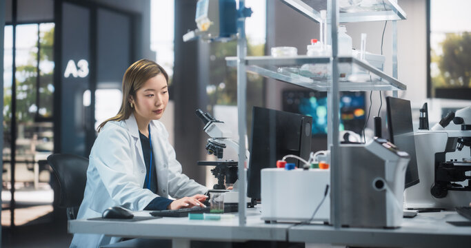 Asina Biotechnologist Studying Genetic Samples Under a Microscope and Researching Data on Computer in Order to Develop New Medical Products. Scientist Working in a Laboratory