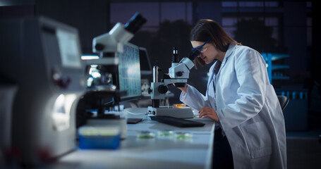 Fototapeta premium Medical Research and Development Laboratory: Hard-Working Female Scientist Using Computer and Looking at a Sample in a Petri Dish Under a Microscope. Advanced Genetic Engineering Lab at Night