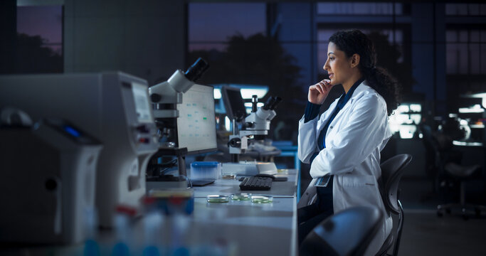 Hispanic Female Scientist Working on a Computer in the Evening. Specialist Using a Gene Sequencing Online Software. Advanced Biotechnology Lab in a Medical Research and Development Center