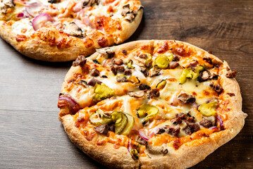 Two delicious pizzas on a wooden surface, one topped with melted cheese, mushrooms, pickles, and savory meat. A perfect treat for pizza lovers, showcasing appetizing colors and textures.