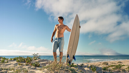 A shirtless man stands on a sandy beach holding a surfboard, enjoying a sunny day by the ocean with clear blue skies.