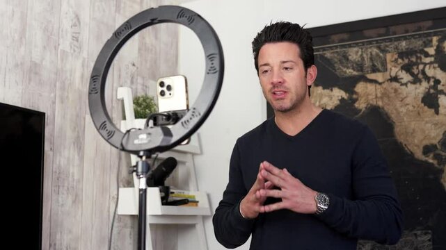 Influencer wearing a black sweater is recording a video blog talking looking at camera, using a smartphone indoors at home.