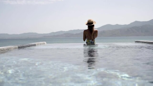 Back view of a woman in swimming costume and sun hat enjoying drink while relaxing on mineral water hot spring in Lesvos Island, Thermi on Yera, Handheld Gimbal shot