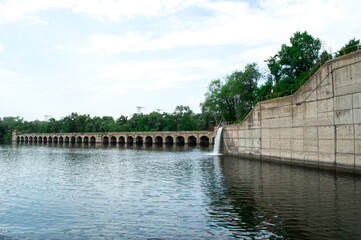 A tranquil scene featuring a dam with arched openings and a waterfall, bordered by verdant trees and reflective water. Suitable for nature and architectural stock photos