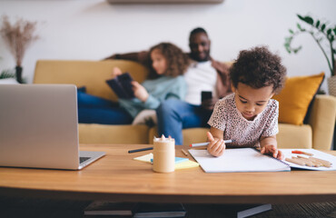 Modern family balancing digital and traditional learning at home interior, Young child focused on drawing while blurred parents relaxes in background in contemporary living space with natural light