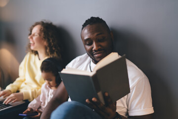 Young African American man enjoying leisure weekend for reading interesting book in home interior, dark skinned hipster guy holding literature poetry in hand and checking text during hobby recreation