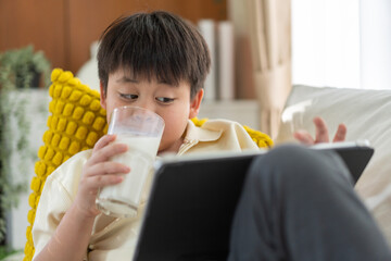 Asian boy holding a glass of milk drinking while playing game on tablet or watching cartoons at...