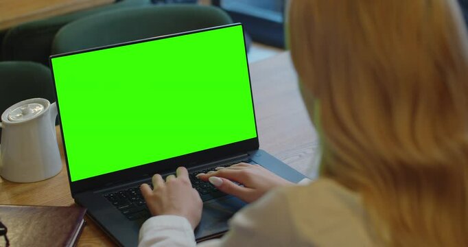 Over the shoulder shot of a business woman working in cafe interior on laptop on desk, looking at green screen. Woman using laptop computer with laptop green screen, sitting at wooden table.