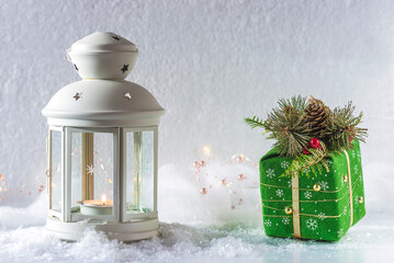 Christmas holiday concept; White lantern with burning candle and green gift box on white snow covered background