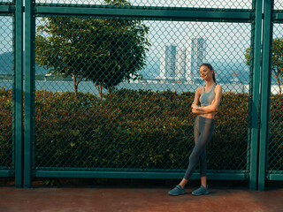 Woman in athletic wear leaning against a fence by a sports court with city skyline in the background