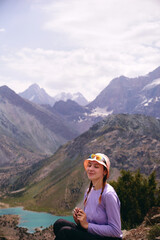A young tourist girl is sitting and smiling against the backdrop of high mountains and a mountain lake. Mountain trekking in summer