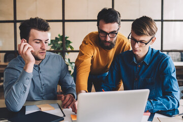 Group of young male coworkers watching webinar online via online application on modern laptop computer, serious skilled men making research for startup project browsing internet on netbook device