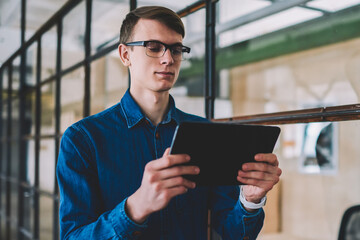 Serious hipster guy in eyeglasses reading information from internet web page using good connection on digital tablet , caucasian man making banking online via application on modern touch pad