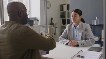 Medium shot of polite Caucasian female visa officer greeting African American male applicant who taking seat and passing documents before interview in embassy