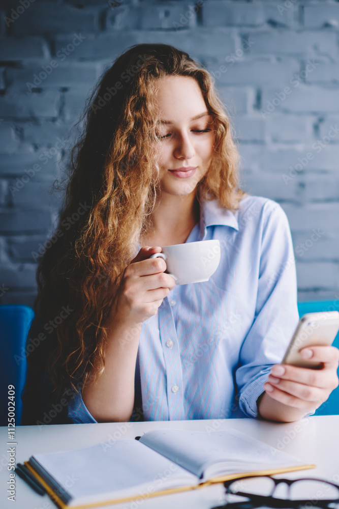 Poster charming female blogger enjoying tea time in cafeteria and sending text messages to followers via sm