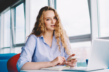 Obraz premium Portrait of tired female freelancer holding smartphone gadget in hand and looking at camera while sitting at desk with laptop computer, young caucasian hipster girl using technology and good internet