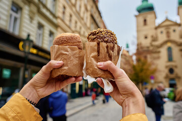 Close-up of woman holding two chimney cakes wrapped in paper with blurred city in background....