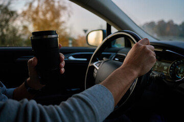 Person drives car on highway road and holding travel mug with hot coffee. Male driver drinks coffee while driving car