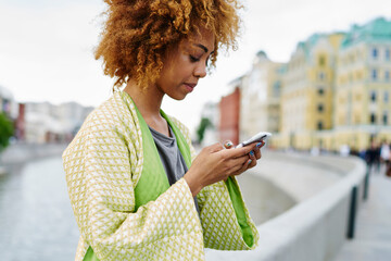 African american young woman with curly hair checking email with notification on smartphone standing in urban setting of architectural town.Dark skinned hipster blogger chatting on telephone