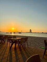 Serene beach at sunset. Tables and chairs on tiled patio. Boats on calm water. Tall building silhouetted against the sky. Long shadows from the setting sun. Tranquil atmosphere with gentle waves.