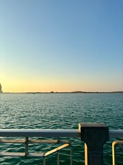 Sunset view from a pier with metal railing. Body of water with gentle waves. Low-lying landmass with trees and small hills in distance. Clear sky gradient from light yellow to deeper blue. Calm and pe