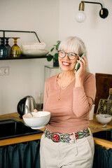 Happy senior woman talking on phone and eating breakfast in kitchen