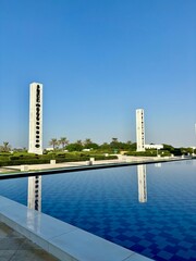 "Two tall, white towers with black circular designs reflect in a large rectangular reflective pool with a tiled bottom. The scene, part of a landscaped park or memorial site, is serene and picturesque