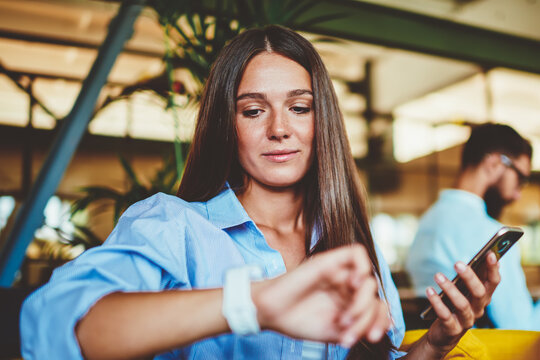 Attractive young woman checking time and notification on electronic wristwatch while spending time with technology on leisure, woman waiting call on cellular phone and reading message on smartwatch