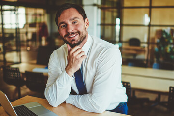 Fototapeta premium Portrait of cheerful male economist in formal wear sitting at desktop with laptop device and smartphone and looking at camera, successful man employee spending time in office and waiting colleague