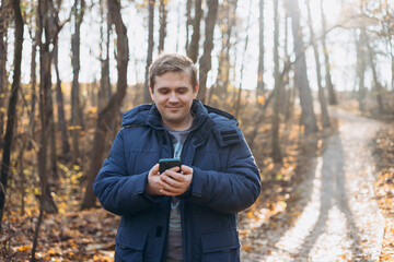 Fototapeta premium Adult 30s man hiker stand outdoors in a natural setting, smile while using his mobile phone outdoors. Hiking, lifestyle, adventure and tourism concept.