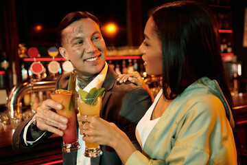 A young couple shares a joyful moment while toasting with colorful drinks in a lively bar.