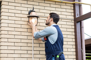 Young handsome electrician standing on ladder and change the light bulb in house facade on the...