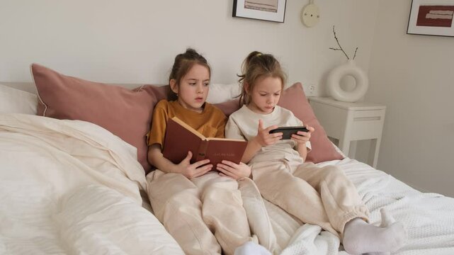 Two twin sisters relax in bed, one engrossed in a book and the other focused on her smartphone. A peaceful, intimate moment capturing sibling interaction and quiet leisure time at home.