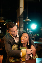 A young multicultural couple enjoys a romantic evening at a lively bar filled with laughter.