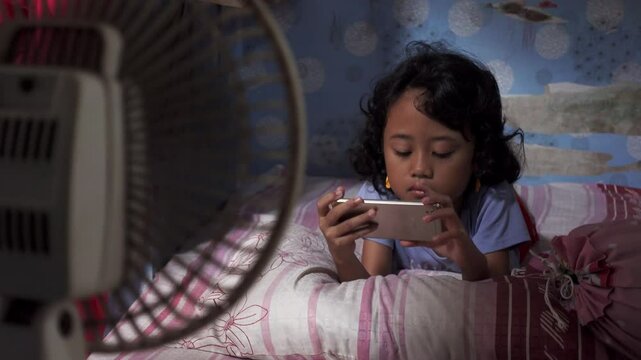 Little girl plays games on smartphone in heat weather in front of the fan. A kid relax on the bed while enjoying the cool breeze from the electric fan during hot weather.
