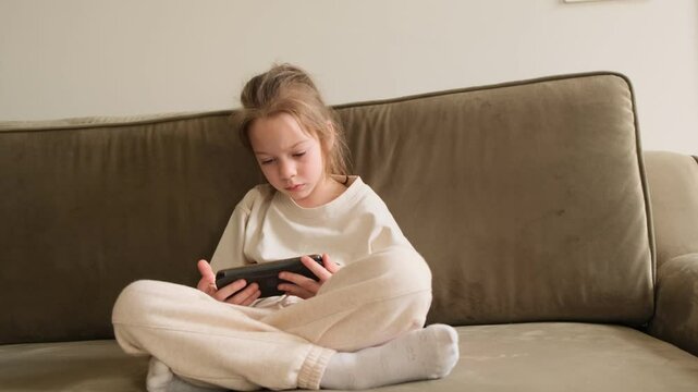 A young girl sits on a couch, engrossed in a smartphone. The focus is on her and the neutral colors of the couch and her clothing. The scene suggests a relaxed, quiet moment at home.