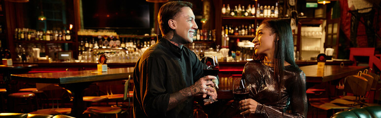 Tattooed man and woman share a romantic night out, holding glasses of wine in a lively bar setting.