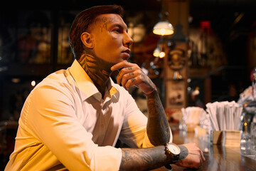 A young man with striking tattoos sits thoughtfully at a bar, embracing the evening vibe.