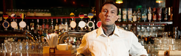 A stylish young man with tattoos relaxes in a bustling bar during an engaging night out.