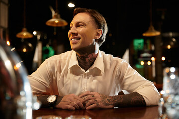 A tattooed young man sits comfortably at the bar, smiling and savoring the evening.