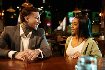 Tattooed man and woman share a warm moment on their date in a vibrant bar setting.
