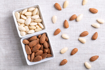 Almonds in square shaped white bowls, on linen fabric. Almonds with seed coats and blanched. Fruits of Prunus amygdalus, syn. Prunus dulcis, ready-to-eat as snack or as ingredient for baking. Photo.
