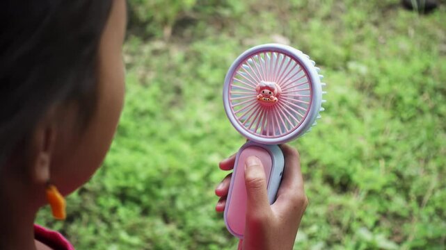 Portable fan held outdoors for quick cooling. Female hand holding mini fan against green grass background.