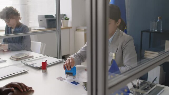 Panning shot of young female Caucasian visa officer approving Schengen visa for African American man during interview in EU consulate